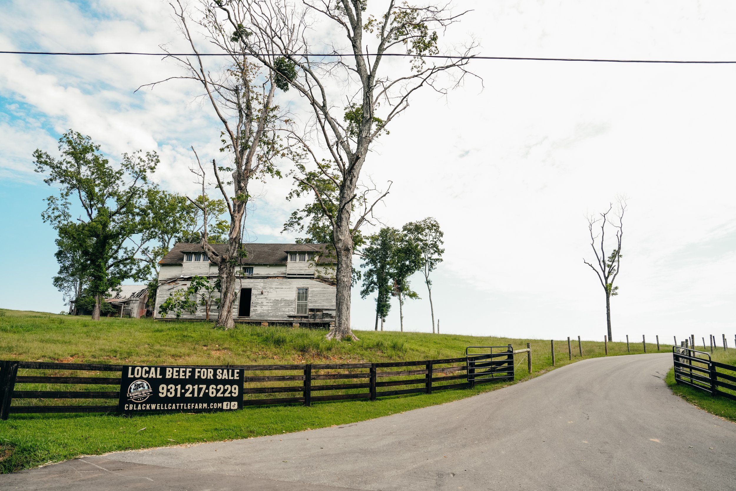 Abandoned farmhouse with a weathered exterior surrounded by trees, featuring a sign advertising local beef for sale and contact information, set against a clear blue sky.