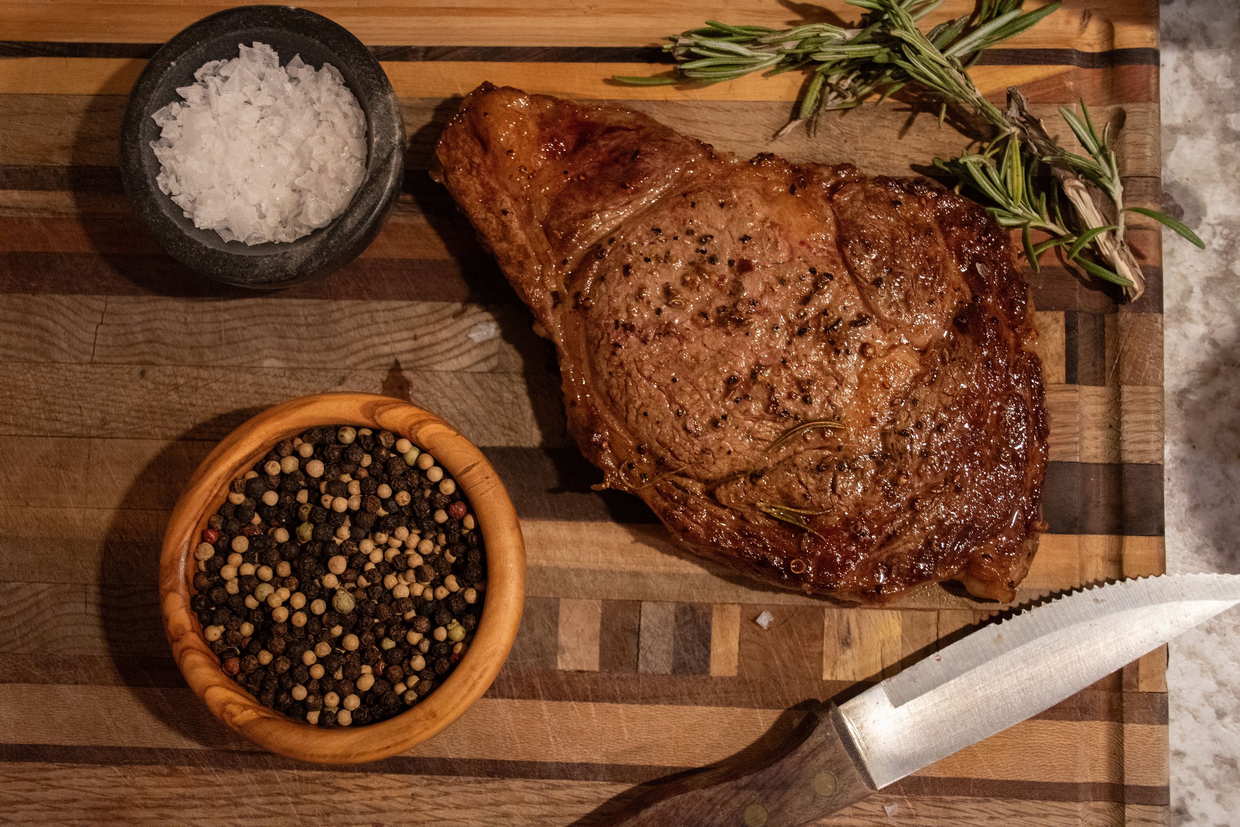 Juicy ribeye steak seasoned with pepper and garnished with fresh rosemary, accompanied by a bowl of coarse sea salt and a wooden bowl of mixed peppercorns, all displayed on a wooden cutting board.